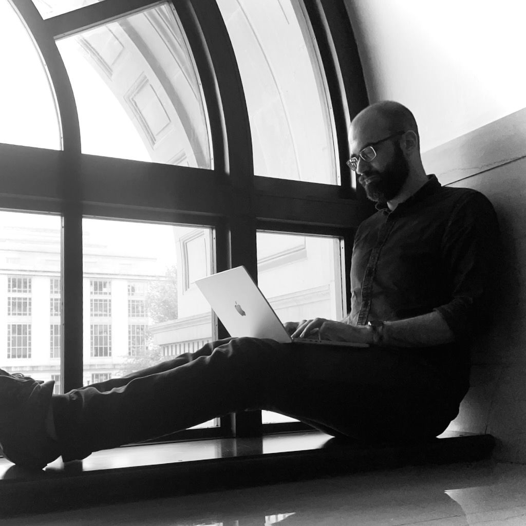 Chris Given types on a laptop while seated on the sill of a large, arched window. He is a white man with a beard and glasses. He wears a dark button-down shirt, slacks, and sneakers. The building is in a neoclassical style, and the National Museum of Natural History is visible outside the window.