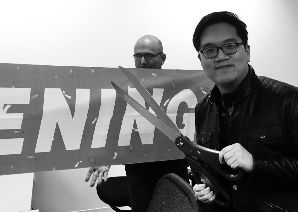 A man stands next to a banner reading 'grand opening' holding a pair of oversized scissors. The scissors are approximately 30 inches (75 centimeters) long. The man wears glasses and a leather jacket, and is of east Asian descent. Another man stands in the background, holding one end of the banner. They are in a windowless room with bare white walls, presumably a nondescript government office building.