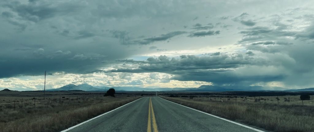 A two-lane road stretches straight to the horizon. The elevation undulates only slightly, and the landscape is mostly brush. There are wire fences to either side of the road and a power line to the left. The sky is filled by dramatic clouds; it has very recently stormed, or it is about to. In the distance, mountains are just coming into view.