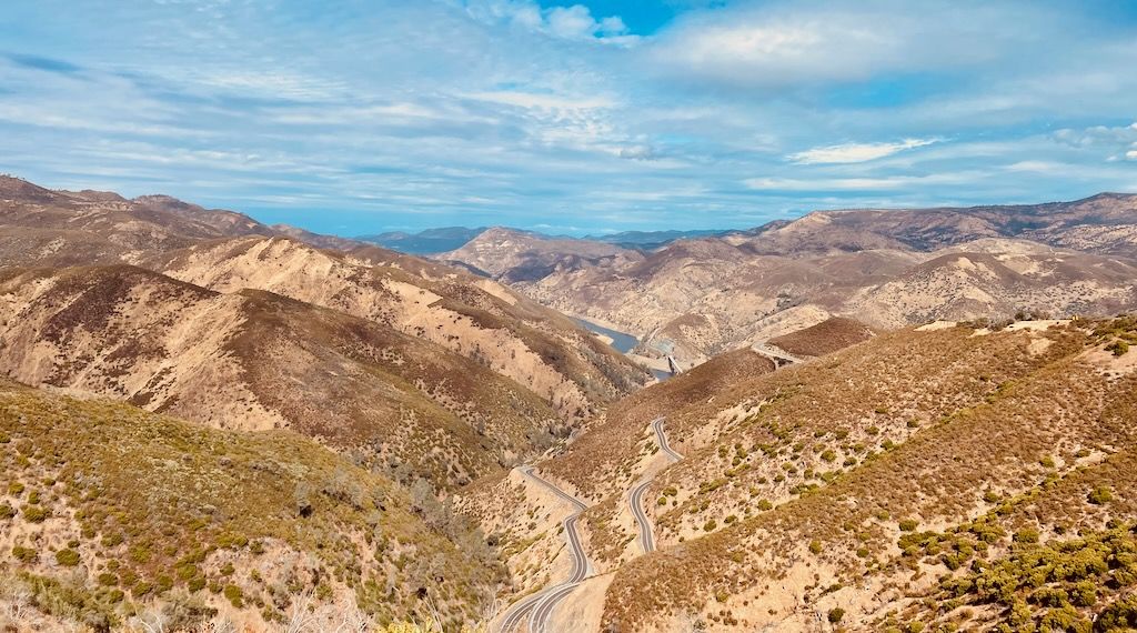 Looking down from a place of high elevation at a road bending sharply through a mountainous terrain. The light brown soil of a relatively arid climate is prevalent, but the mountains are dotted with low brownish-green trees. In the distance, a river leads toward a valley.