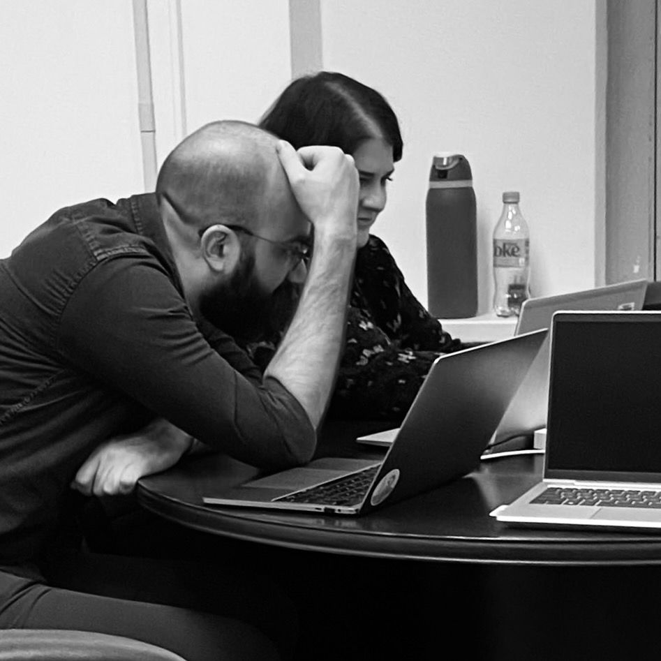 Chris Given and Bridget Roberts sit next to each other, scrutinizing a laptop screen. They sit at a round table in a nondescript government office. Chris is a white man with glasses and a beard wearing a dark button-down shirt. He is hunched over to be close to the screen, one hand resting on his forehead, the other gripping the table nervously. Bridget is a white woman with dark hair wearing a dark, patterned blouse. Of the two, she appears decidedly more placid. A large water bottle and an empty Diet Coke bottle sit on a shelf near her. There are at least three laptops cluttering the table, two PCs and one Mac; the Mac is adorned by a circular sticker with an abstract illustration of a flamingo and a fern in front of an ocean.